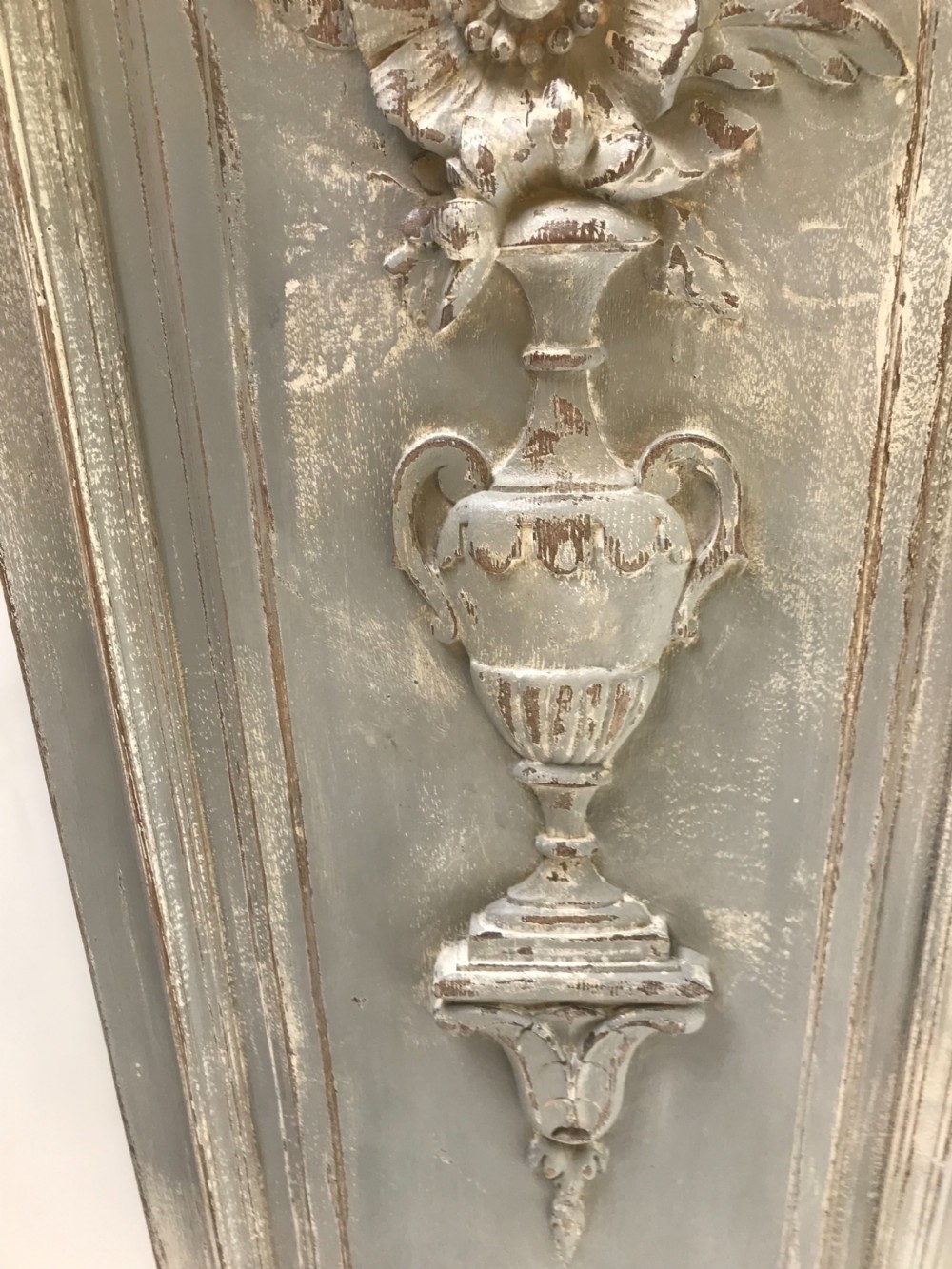 A vertical painted grey wood panel featuring an arched frame encircling a hand-carved classical urn and floral relief. View V