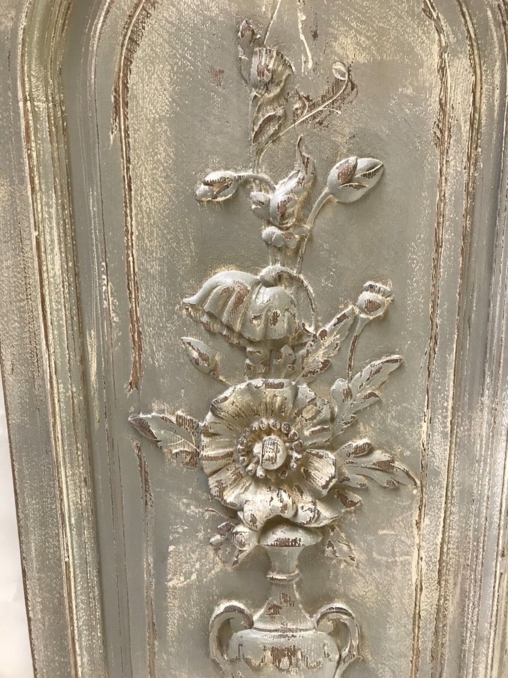 A vertical painted grey wood panel featuring an arched frame encircling a hand-carved classical urn and floral relief. View VI