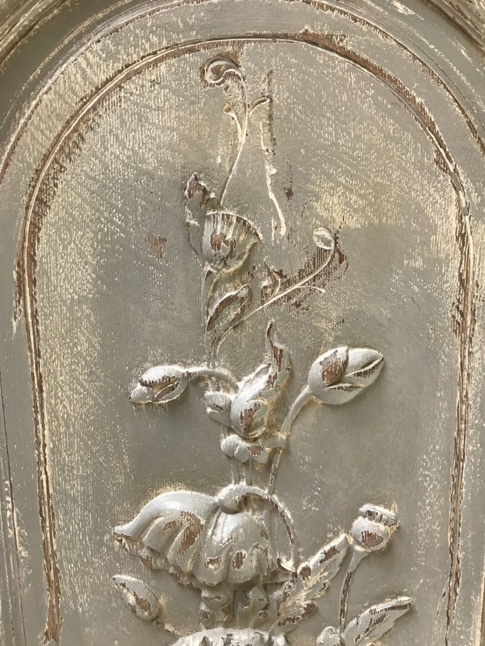 A vertical painted grey wood panel featuring an arched frame encircling a hand-carved classical urn and floral relief. View VII