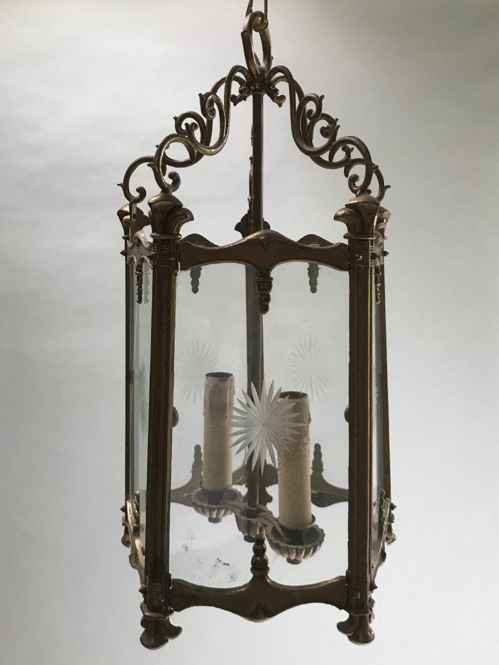 An antique French brass hanging lantern featuring scrolled top details, internal candle-style fittings, and clear glass panels decorated with etched starburst patterns.