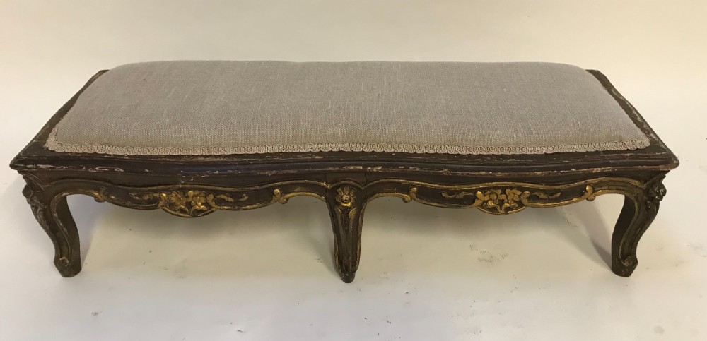 An elongated low rectangular French style footstool featuring a dark giltwood frame with carved floral aprons and six cabriole legs, topped with light grey linen upholstery.