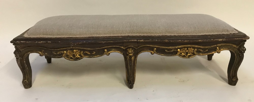 An elongated low rectangular French style footstool featuring a dark giltwood frame with carved floral aprons and six cabriole legs, topped with light grey linen upholstery. View IV