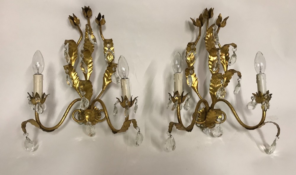 A pair of gilded metal two-arm wall sconces decorated with leaf forms, tulip-shaped flower candle sockets, and suspended clear crystal drops.
