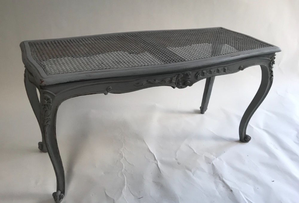 A rectangular grey painted French stool featuring a hand-woven cane top, floral carved apron, and cabriole legs.