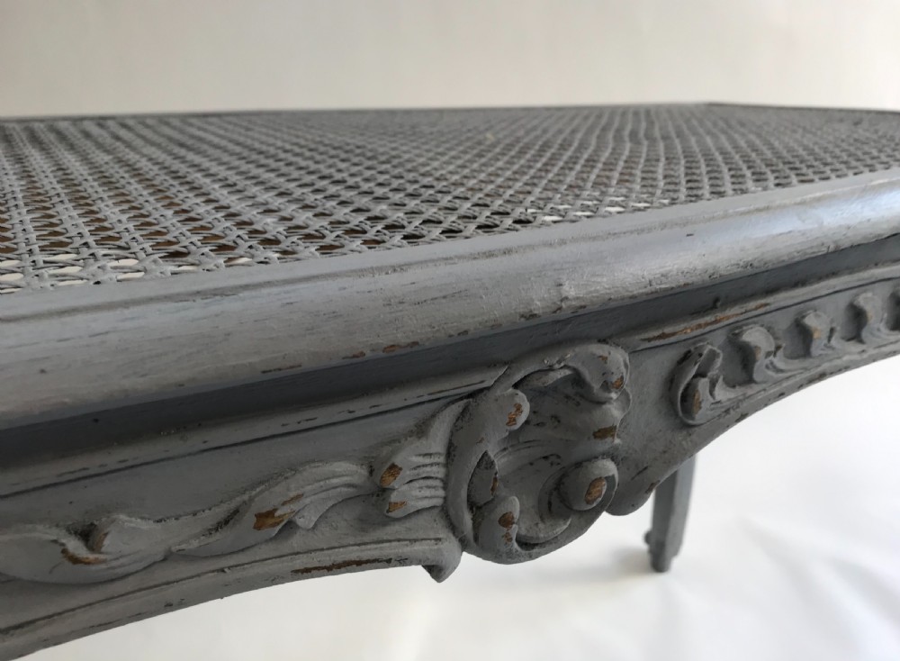 A rectangular grey painted French stool featuring a hand-woven cane top, floral carved apron, and cabriole legs. View II