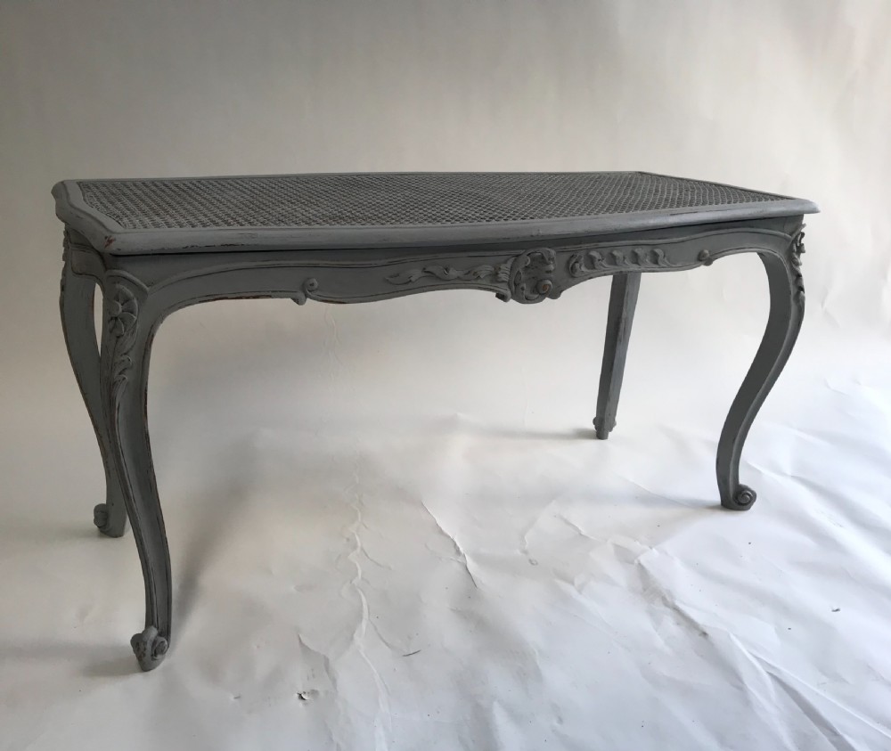A rectangular grey painted French stool featuring a hand-woven cane top, floral carved apron, and cabriole legs. View IV