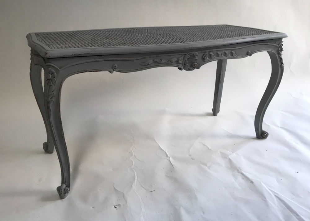 A rectangular grey painted French stool featuring a hand-woven cane top, floral carved apron, and cabriole legs. View V