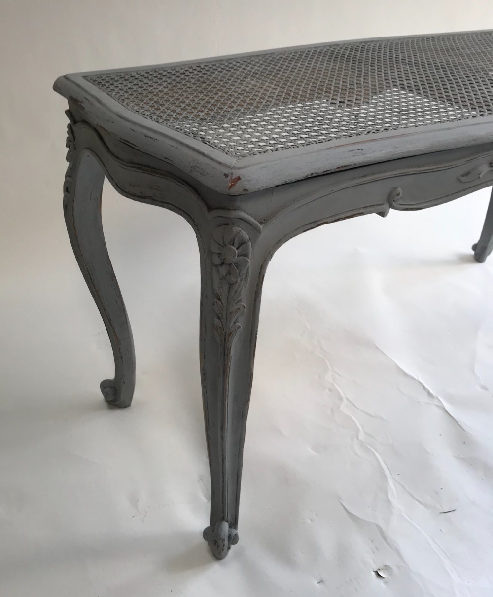 A rectangular grey painted French stool featuring a hand-woven cane top, floral carved apron, and cabriole legs. View VII