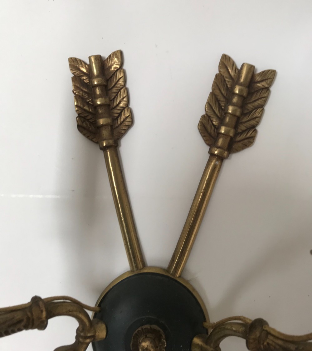 A pair of French brass twin-arm wall lights with a crossed arrow motif and dark circular backplates. View VI