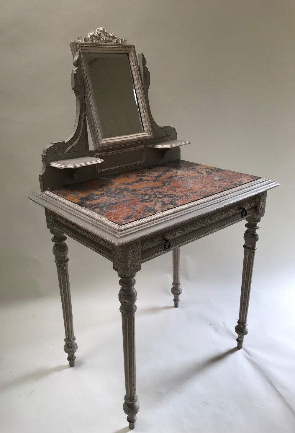 A French painted grey dressing table featuring a variegated reddish-brown marble top, an adjustable central mirror with a carved crest, two small side shelves, and tapered fluted legs.