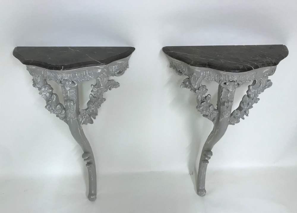 A pair of grey painted carved wood wall consoles featuring floral scroll legs and fitted black veined marble tops.
