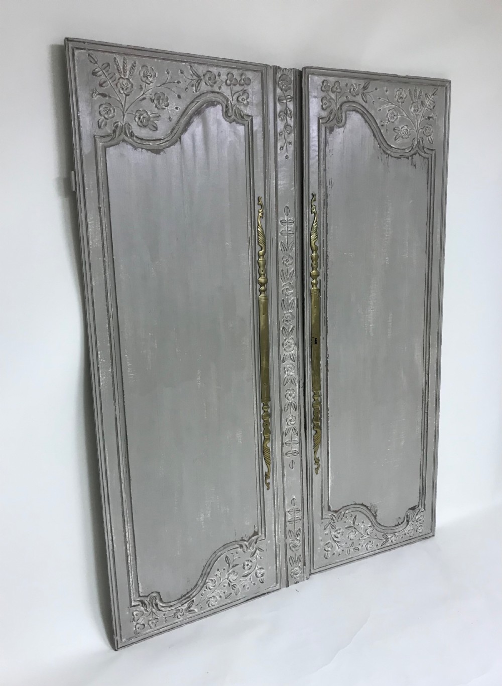 A pair of grey painted antique French doors featuring hand-carved floral panels and long brass handles.