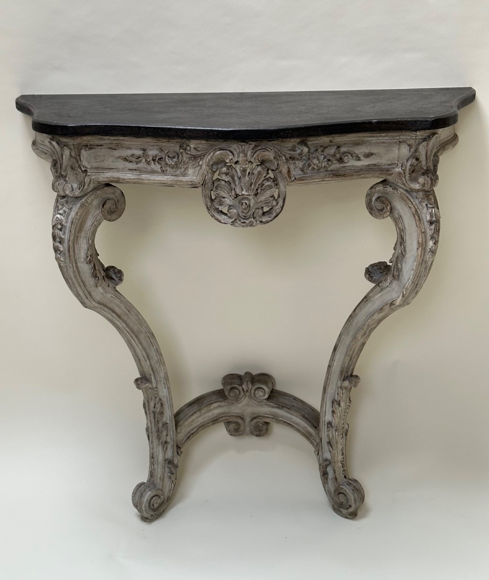 A carved French style console table featuring a distressed grey painted frame, curved cabriole legs, central shell motif, and a dark marble top.
