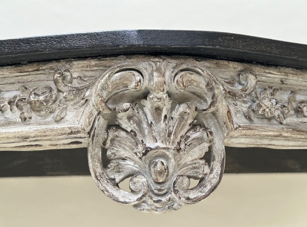A carved French style console table featuring a distressed grey painted frame, curved cabriole legs, central shell motif, and a dark marble top. View II