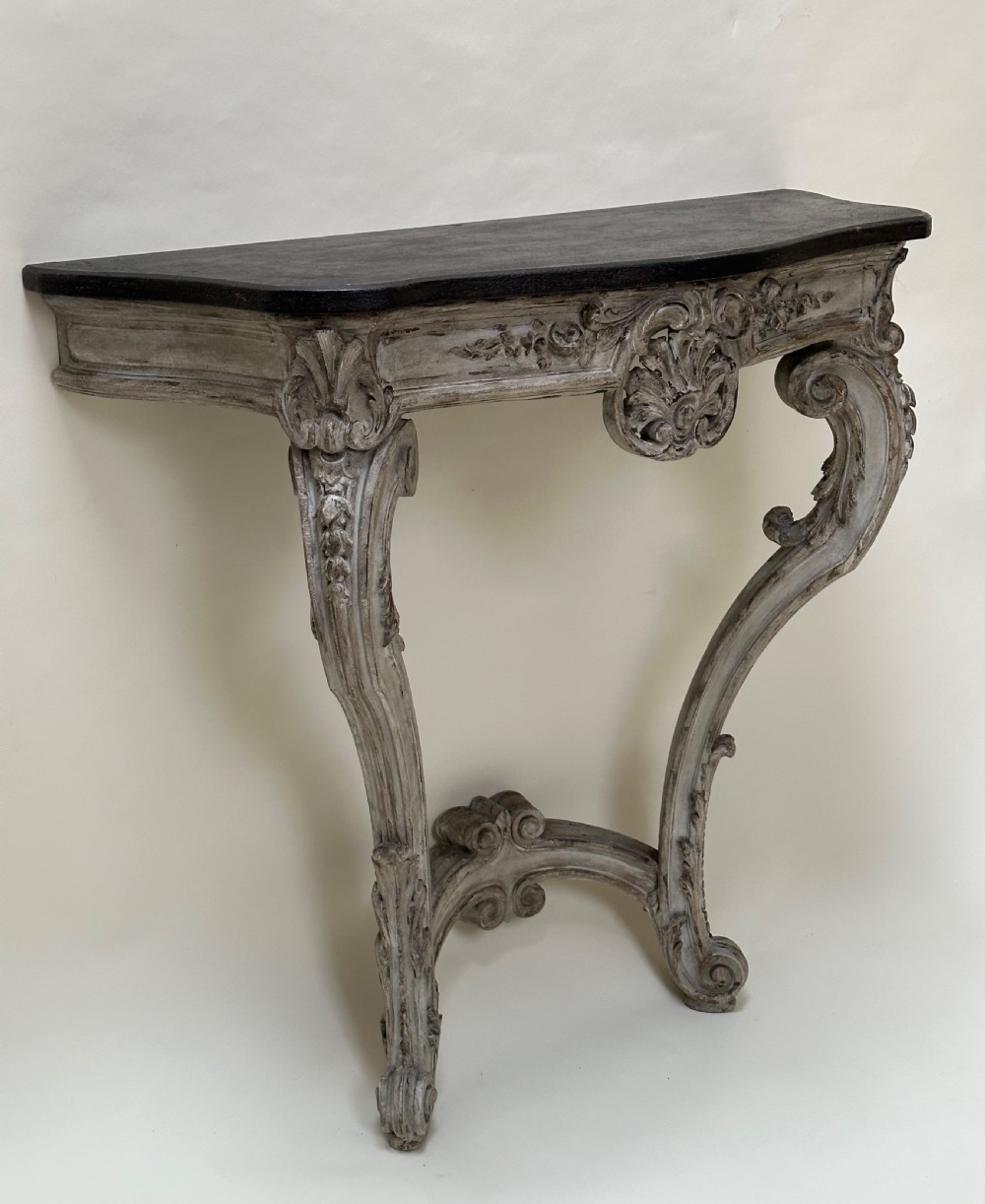 A carved French style console table featuring a distressed grey painted frame, curved cabriole legs, central shell motif, and a dark marble top. View III