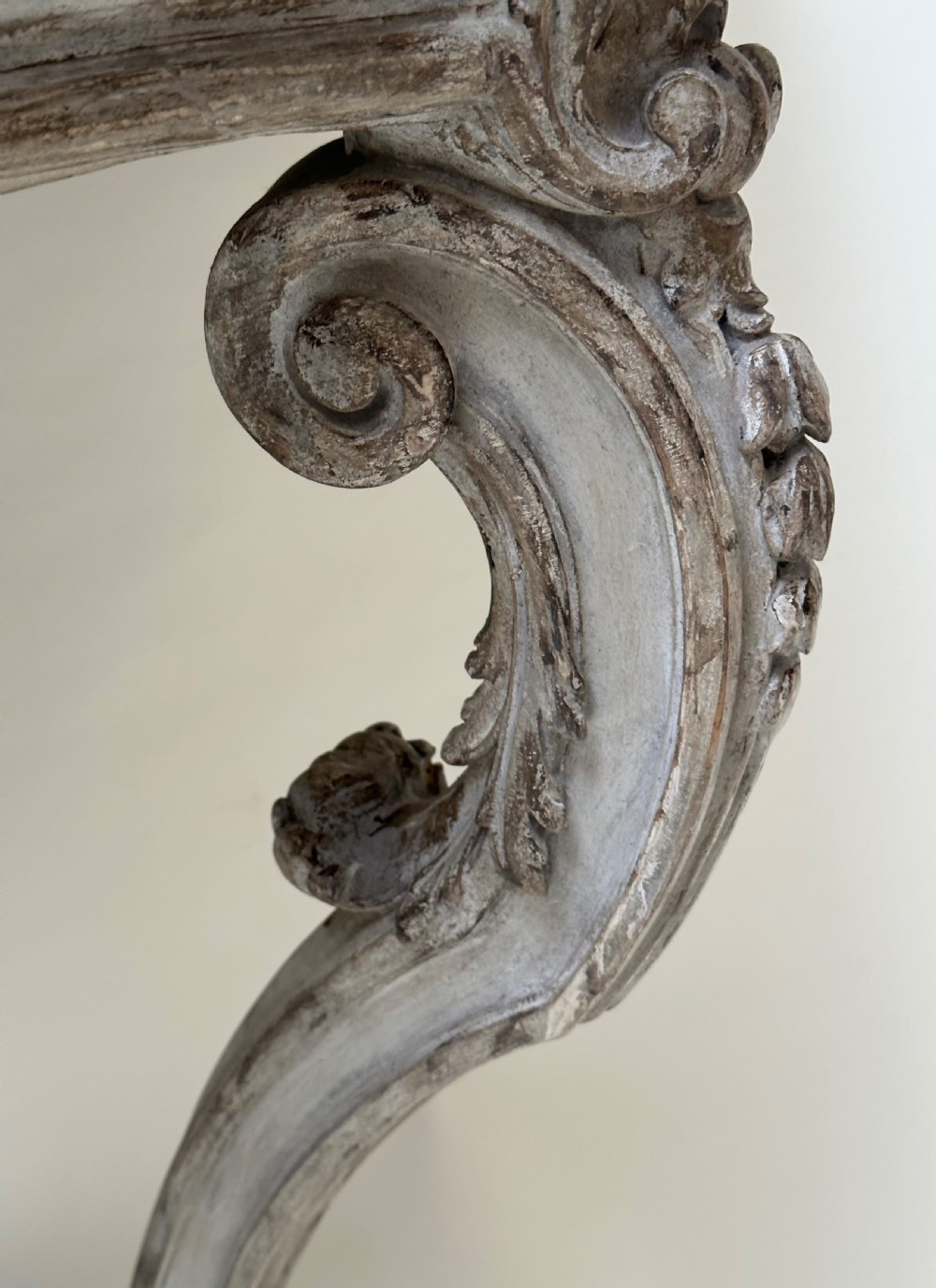 A carved French style console table featuring a distressed grey painted frame, curved cabriole legs, central shell motif, and a dark marble top. View IV