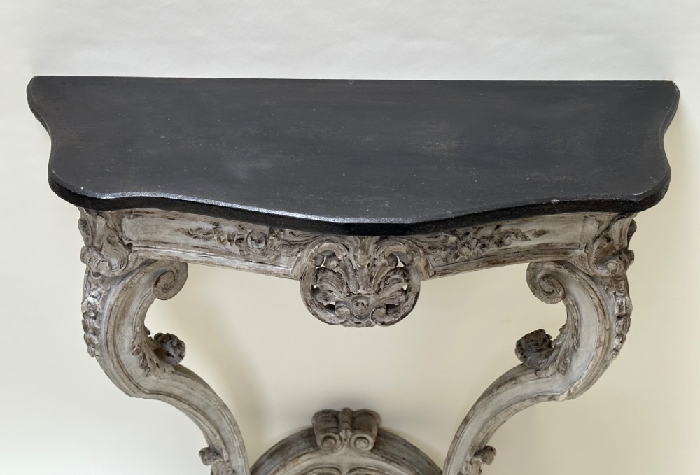 A carved French style console table featuring a distressed grey painted frame, curved cabriole legs, central shell motif, and a dark marble top. View VII