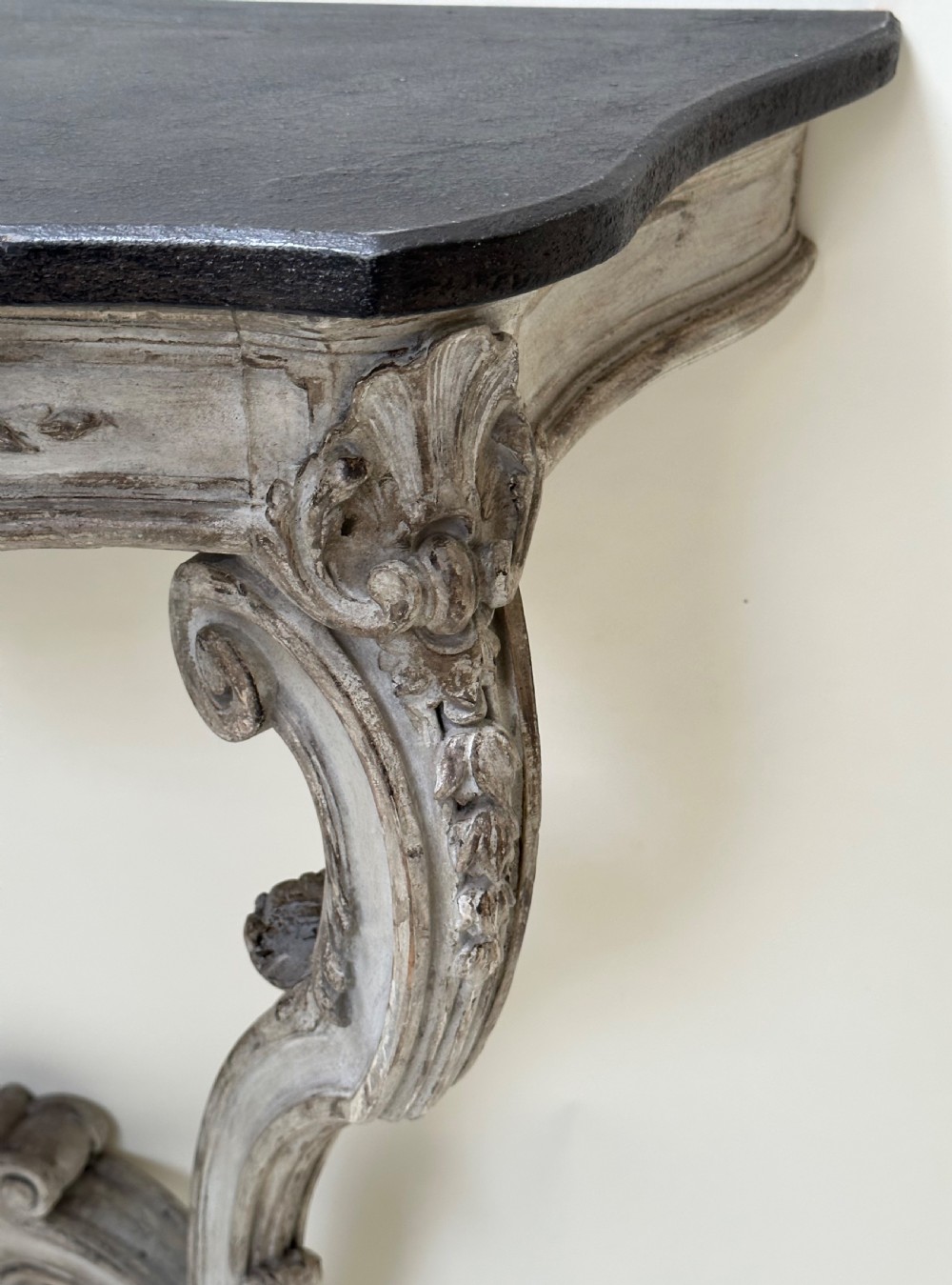 A carved French style console table featuring a distressed grey painted frame, curved cabriole legs, central shell motif, and a dark marble top. View VIII