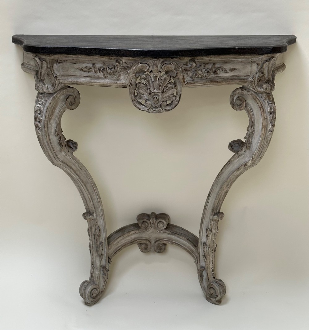 A carved French style console table featuring a distressed grey painted frame, curved cabriole legs, central shell motif, and a dark marble top. View IX