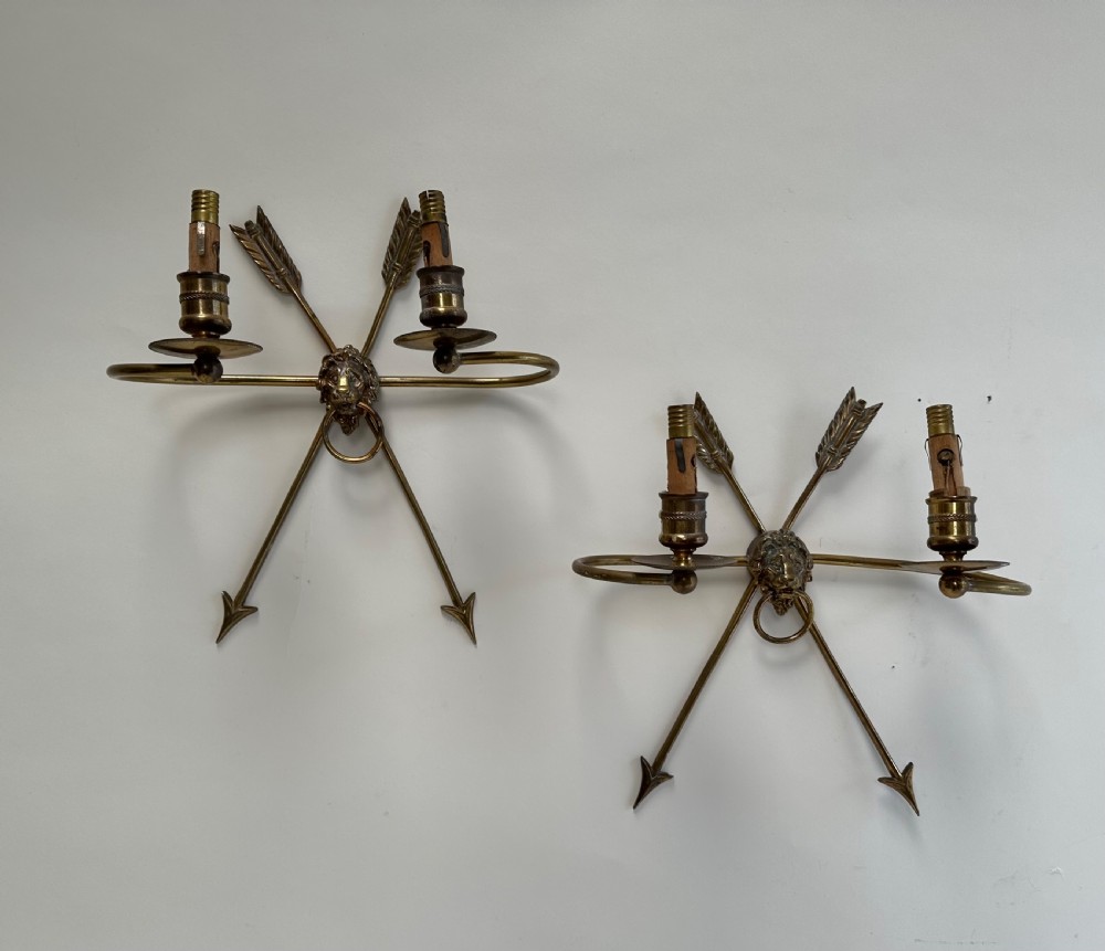A pair of French cast brass wall sconces featuring crossed arrow backplates centred by lion masks with ring handles and two curving candle arms.