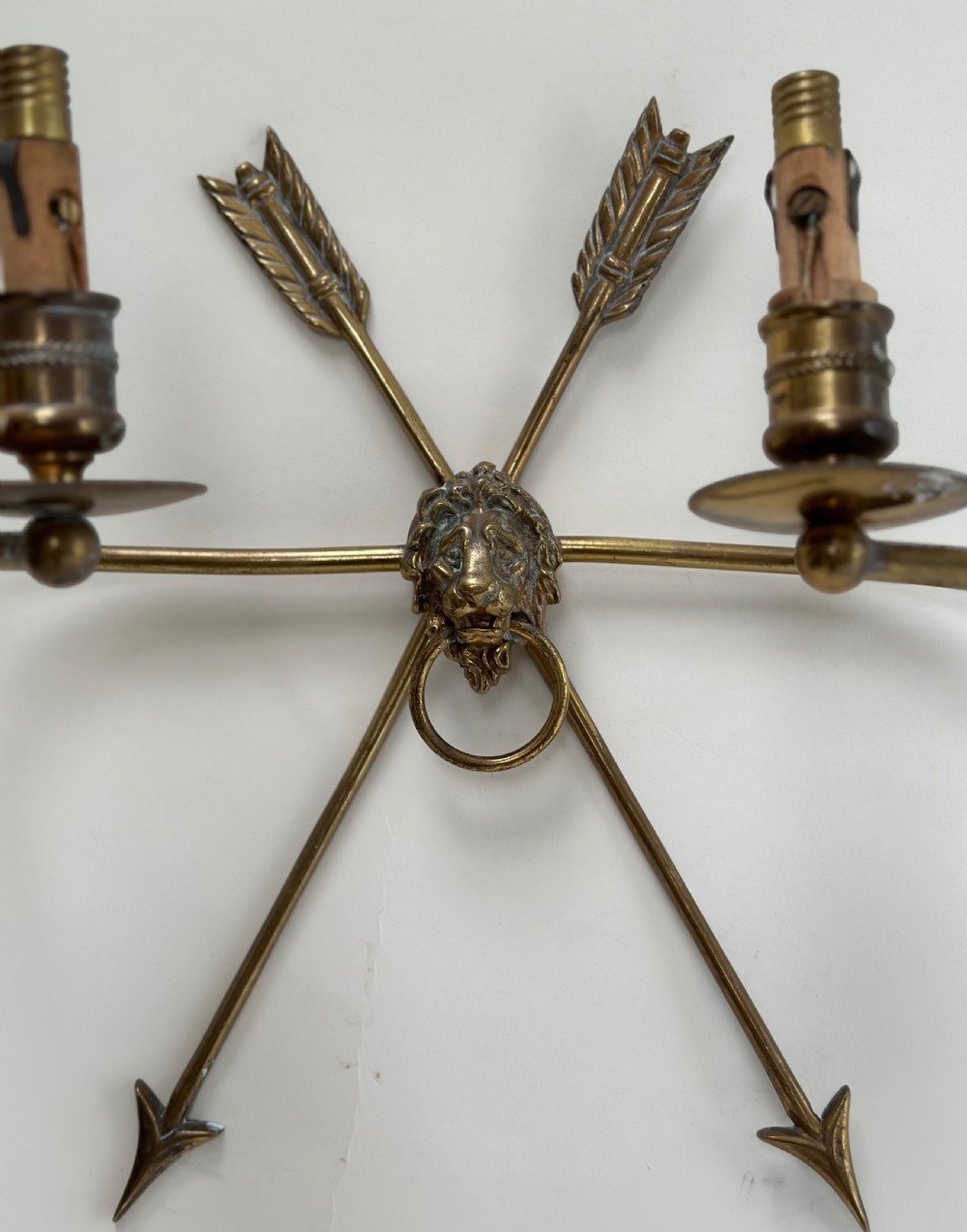 A pair of French cast brass wall sconces featuring crossed arrow backplates centred by lion masks with ring handles and two curving candle arms. View II