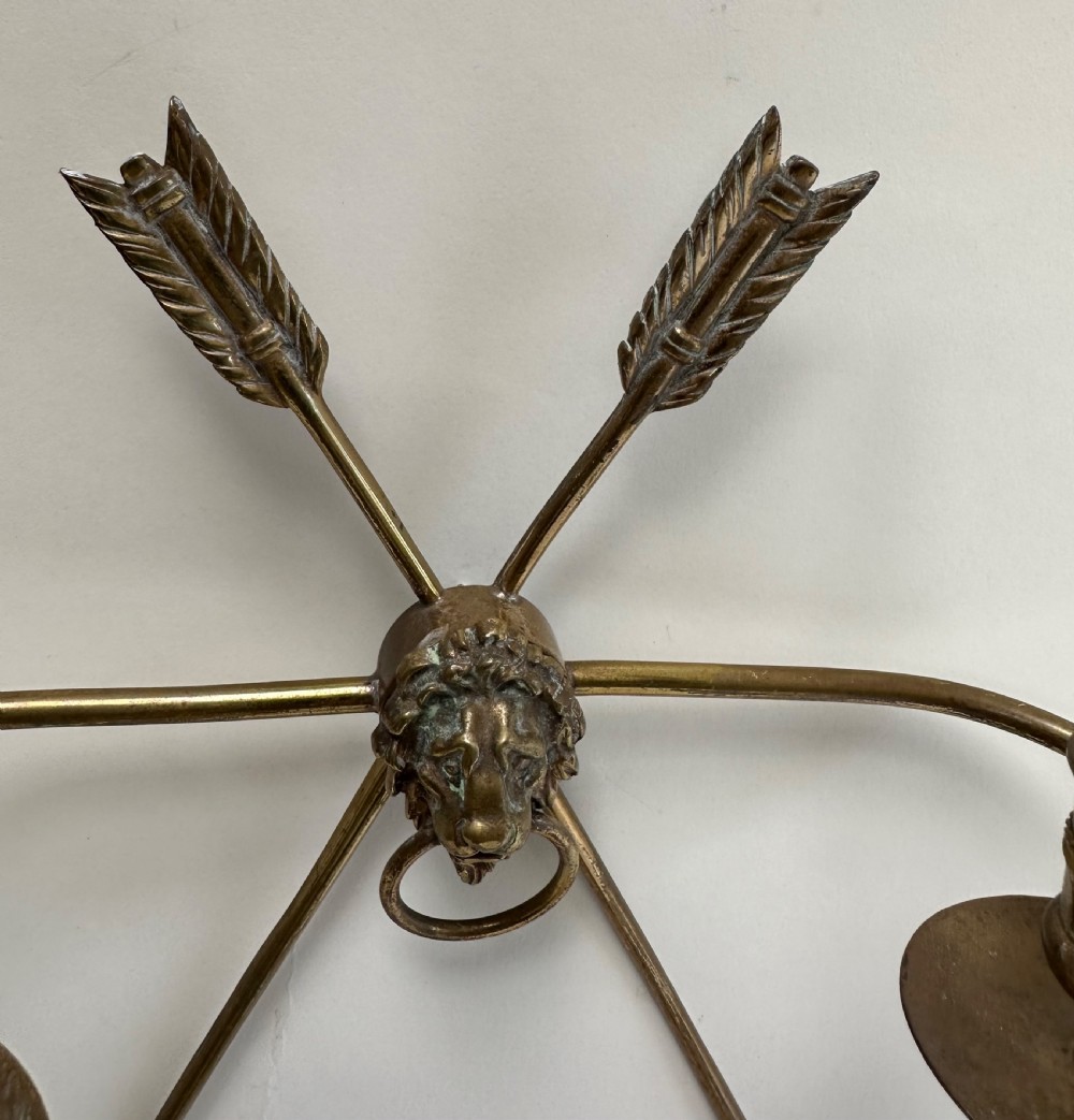 A pair of French cast brass wall sconces featuring crossed arrow backplates centred by lion masks with ring handles and two curving candle arms. View V