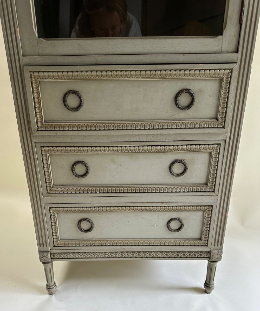 A tall grey-painted French provincial cabinet featuring an upper open compartment, a central glazed glass door, three lower drawers with ring handles, and fluted pilasters. View III