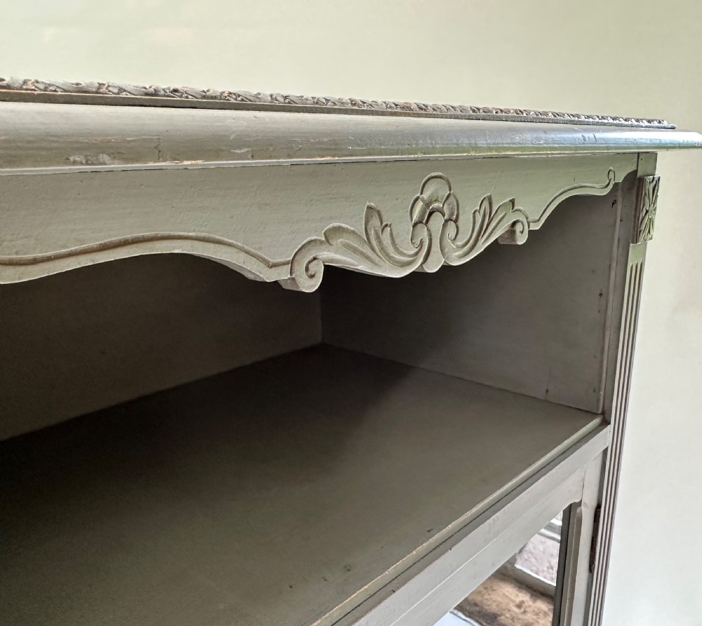 A tall grey-painted French provincial cabinet featuring an upper open compartment, a central glazed glass door, three lower drawers with ring handles, and fluted pilasters. View VIII