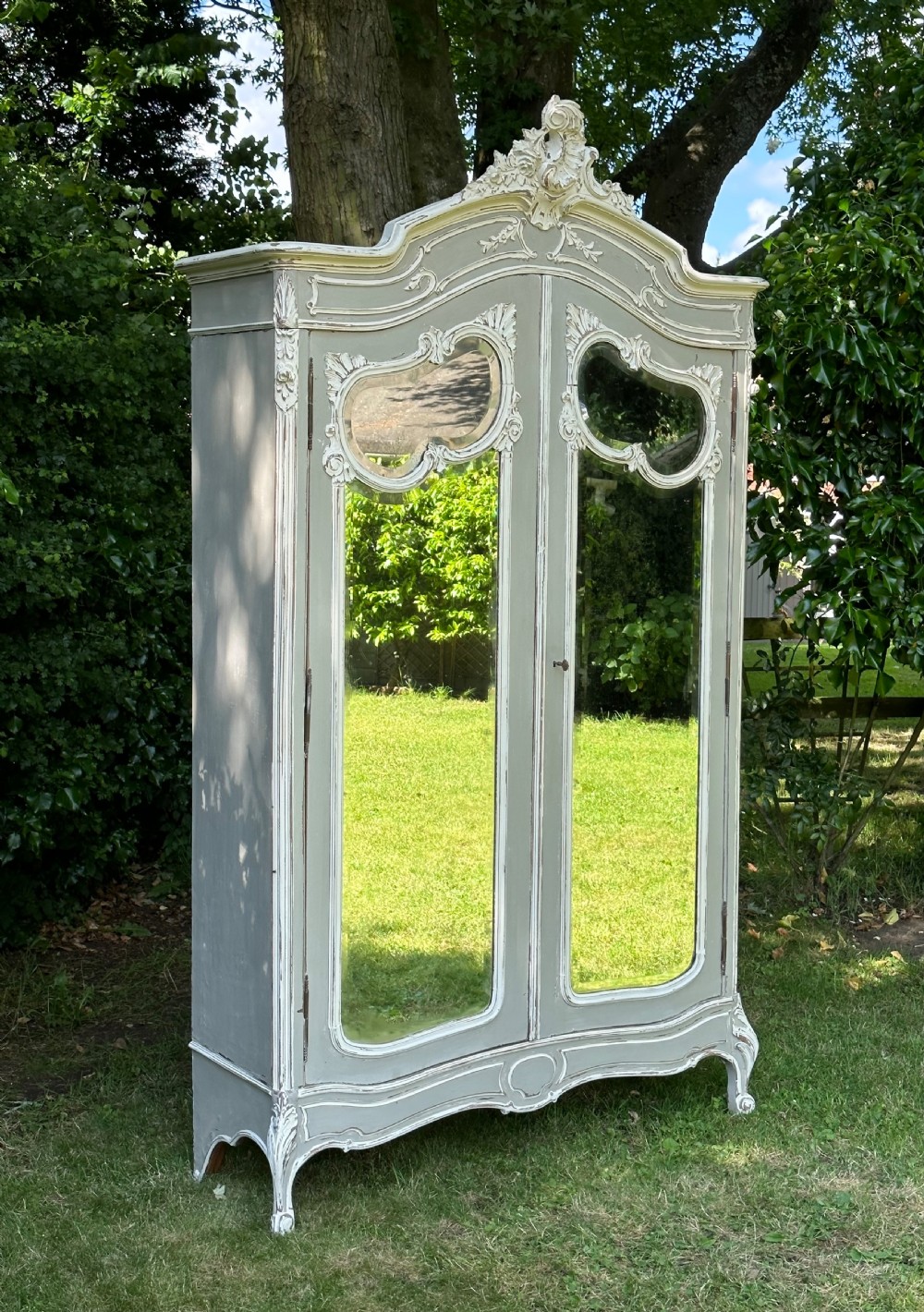 A tall grey and off-white painted French armoire featuring arched mirrored double doors, carved cresting at the top, and short cabriole legs.