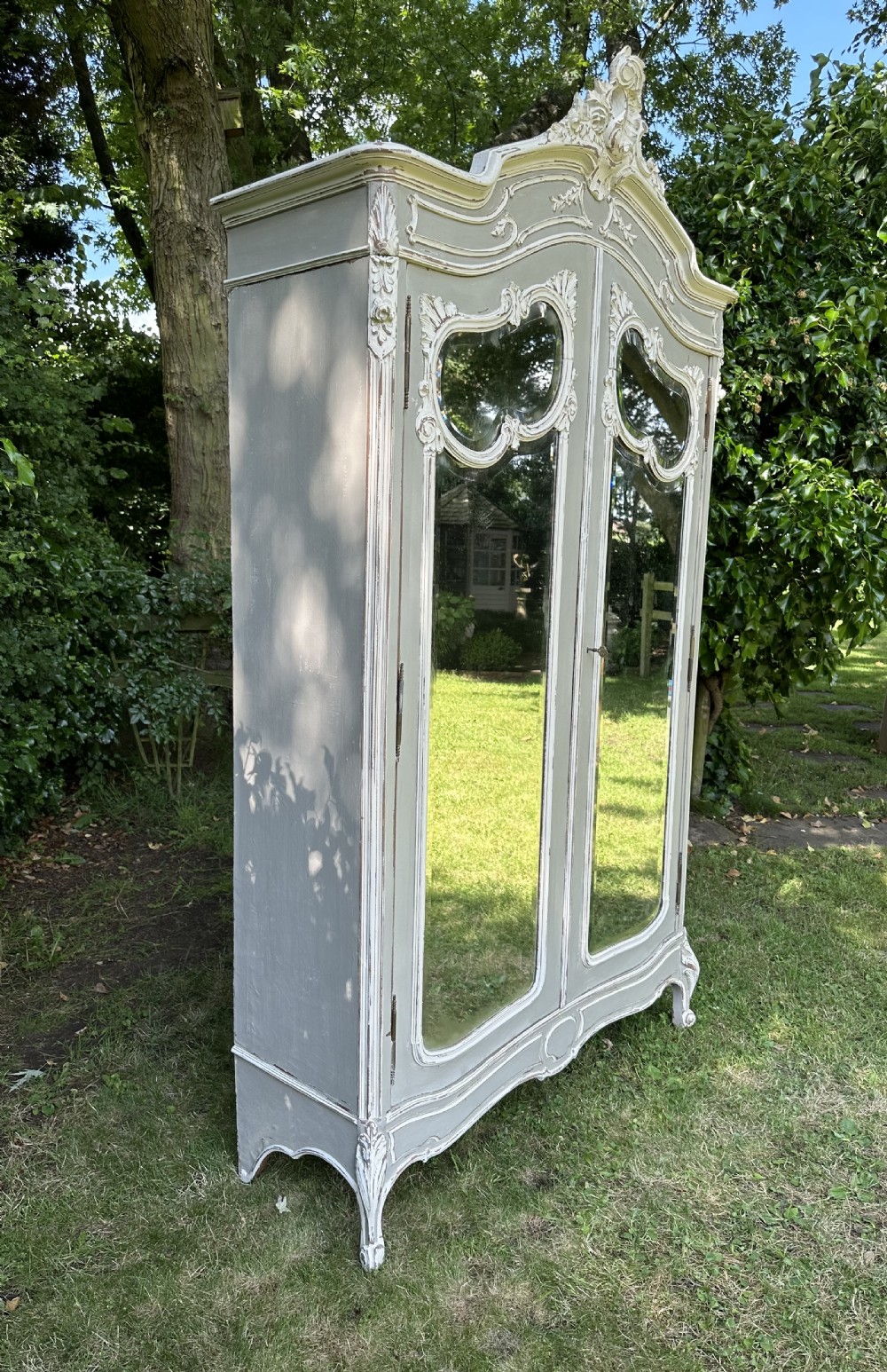 A tall grey and off-white painted French armoire featuring arched mirrored double doors, carved cresting at the top, and short cabriole legs. View VII