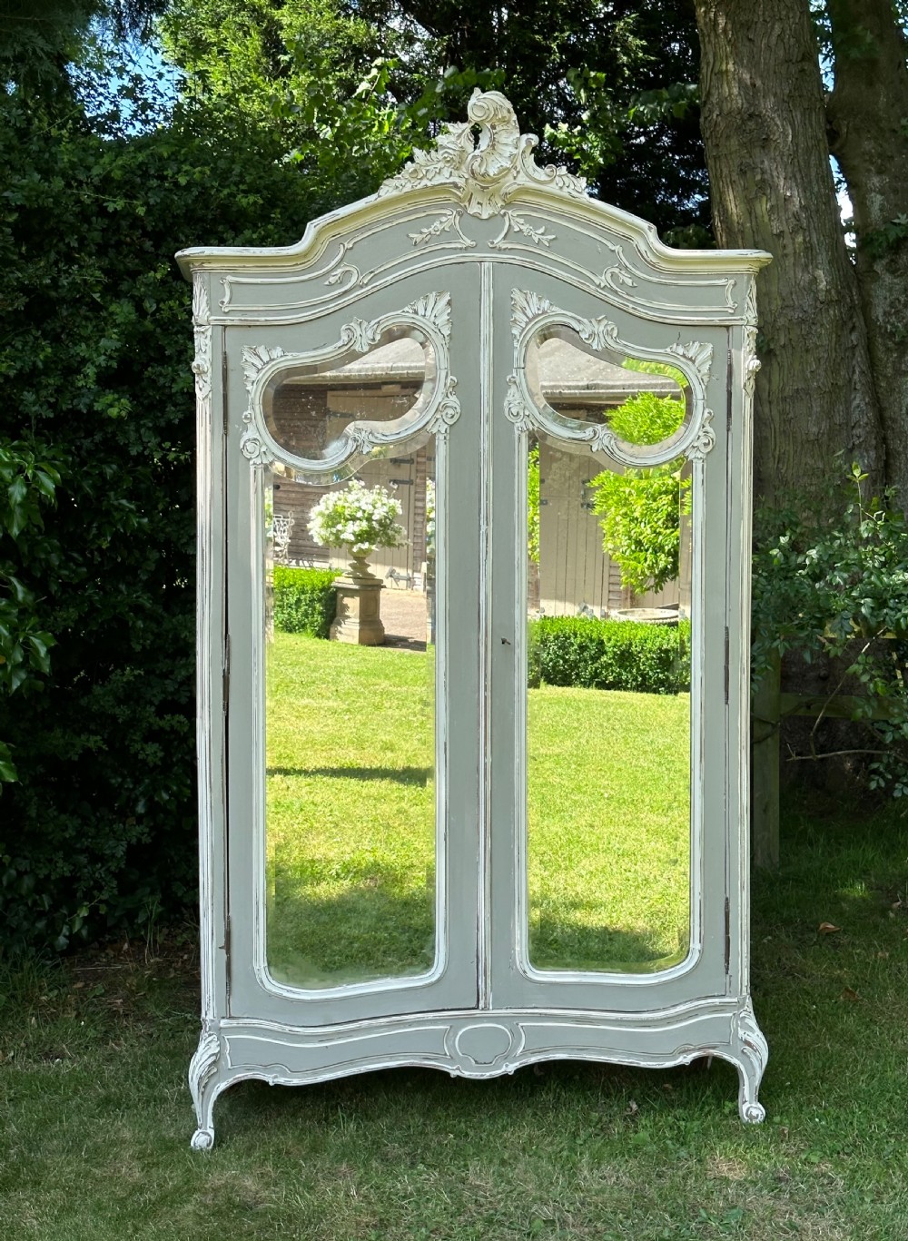 A tall grey and off-white painted French armoire featuring arched mirrored double doors, carved cresting at the top, and short cabriole legs. View X