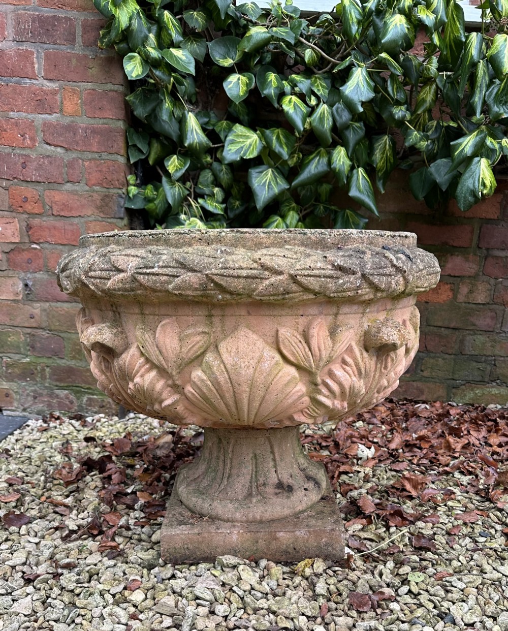 A weathered terracotta garden urn featuring carved relief shell and leaf motifs on a flared pedestal base with a square plinth.
