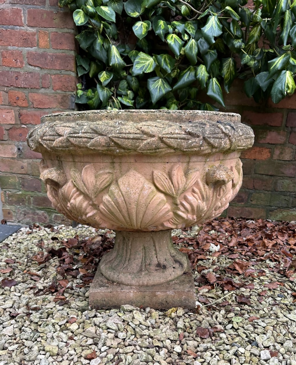 A weathered terracotta garden urn featuring carved relief shell and leaf motifs on a flared pedestal base with a square plinth. View II