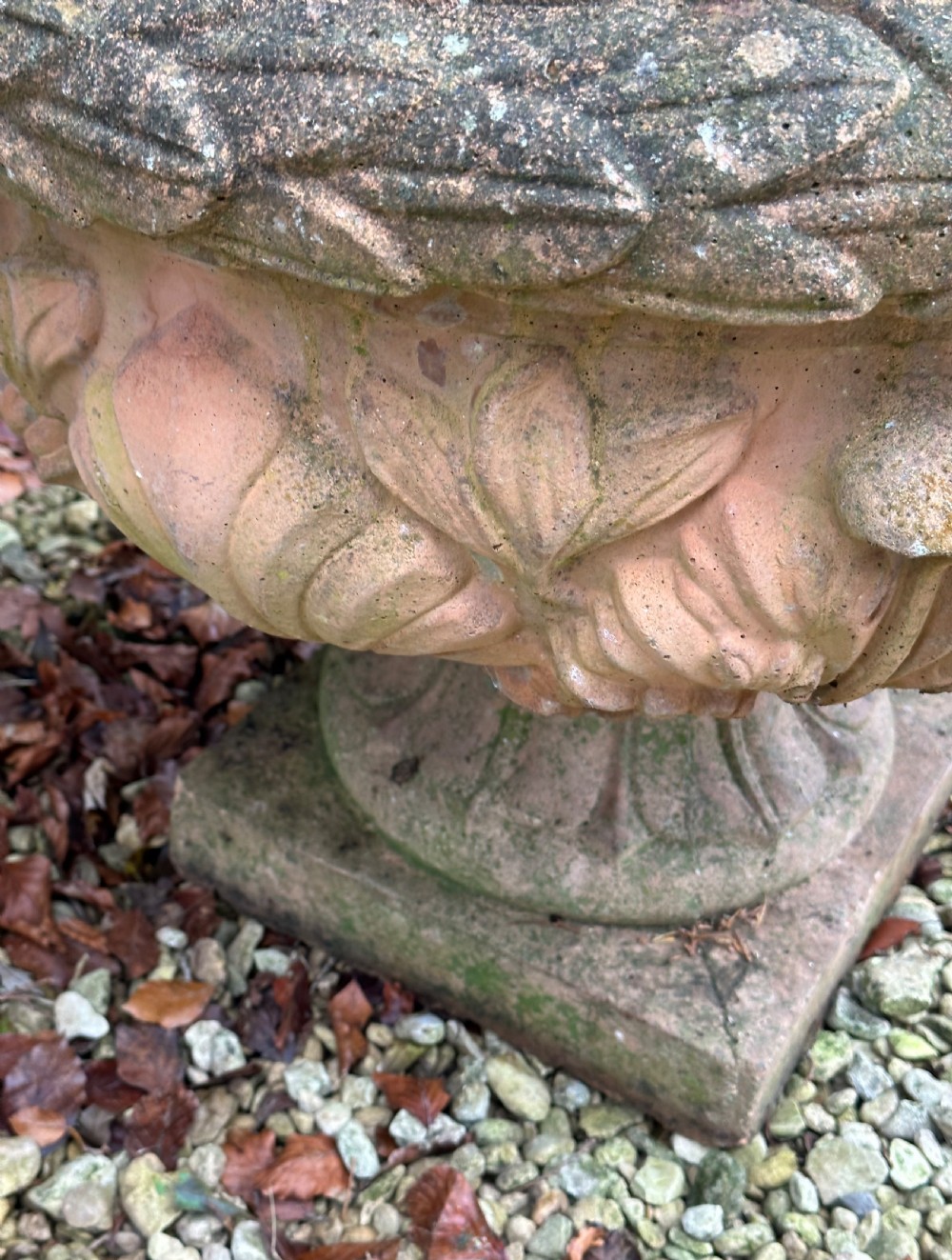 A weathered terracotta garden urn featuring carved relief shell and leaf motifs on a flared pedestal base with a square plinth. View III