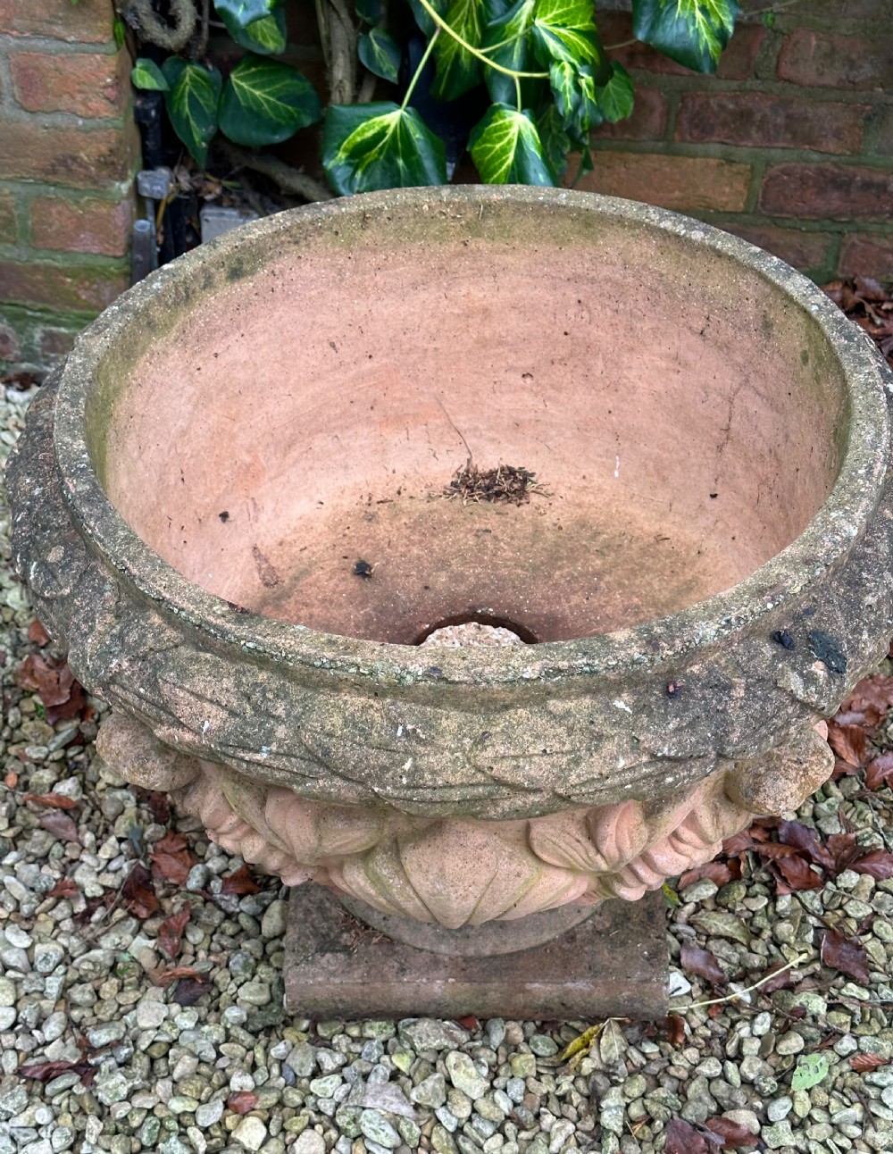 A weathered terracotta garden urn featuring carved relief shell and leaf motifs on a flared pedestal base with a square plinth. View V