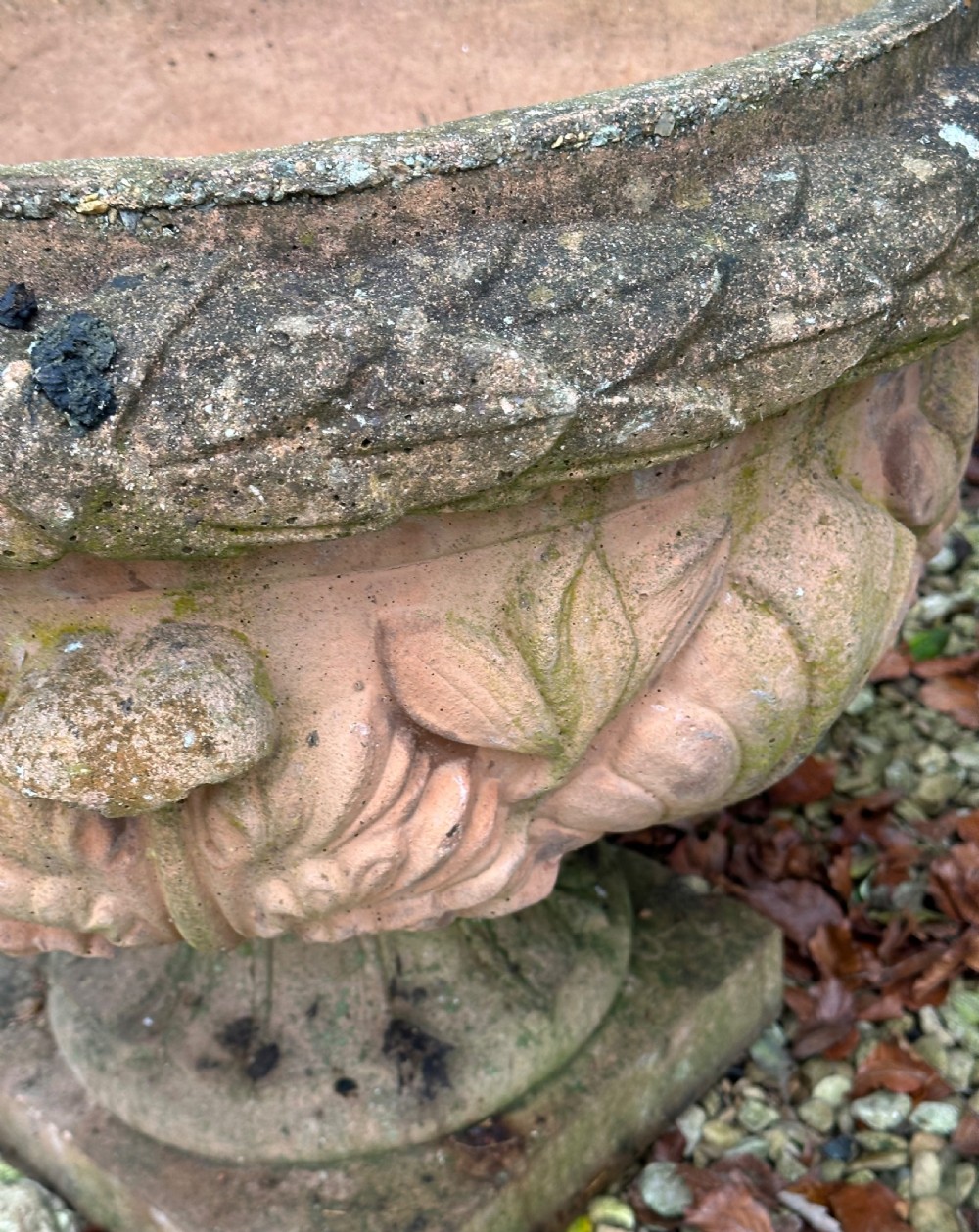 A weathered terracotta garden urn featuring carved relief shell and leaf motifs on a flared pedestal base with a square plinth. View VI
