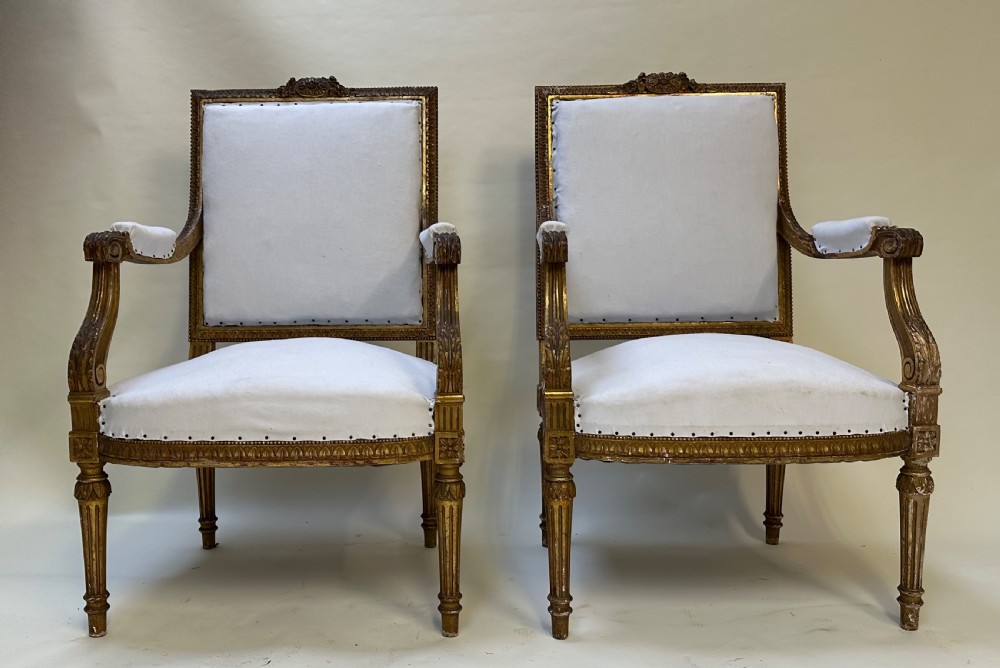 A pair of French Louis XVI style giltwood armchairs with carved square backs, padded armrests, and white fabric upholstery.