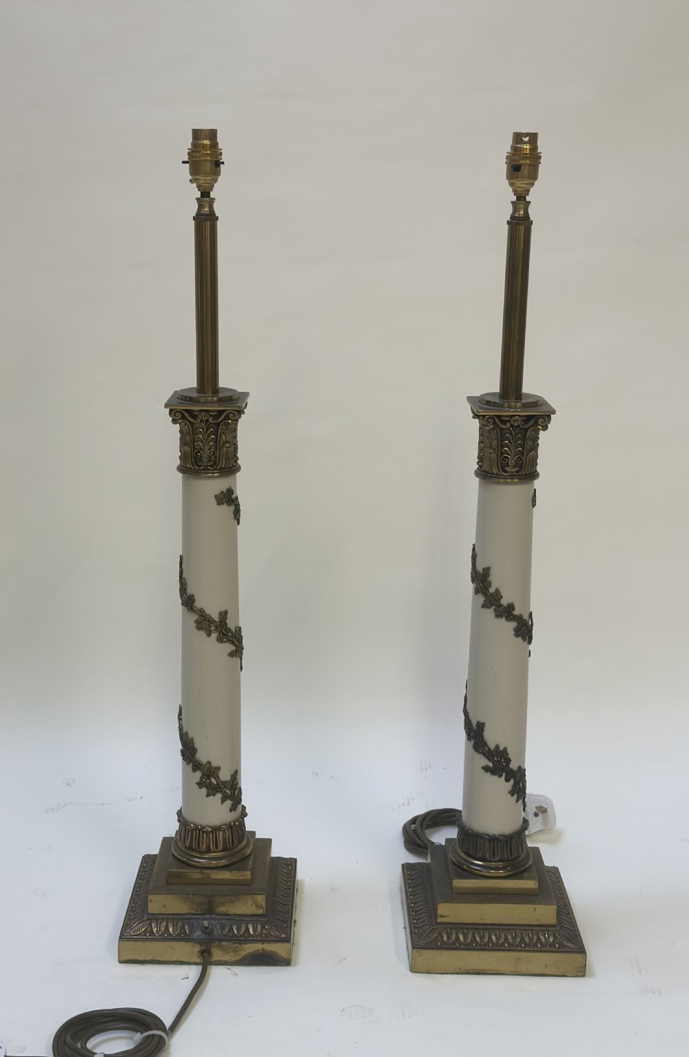 A pair of tall Neoclassical table lamps featuring cream shafts with brass garland detail and brass Corinthian capitals on stepped bases. View V