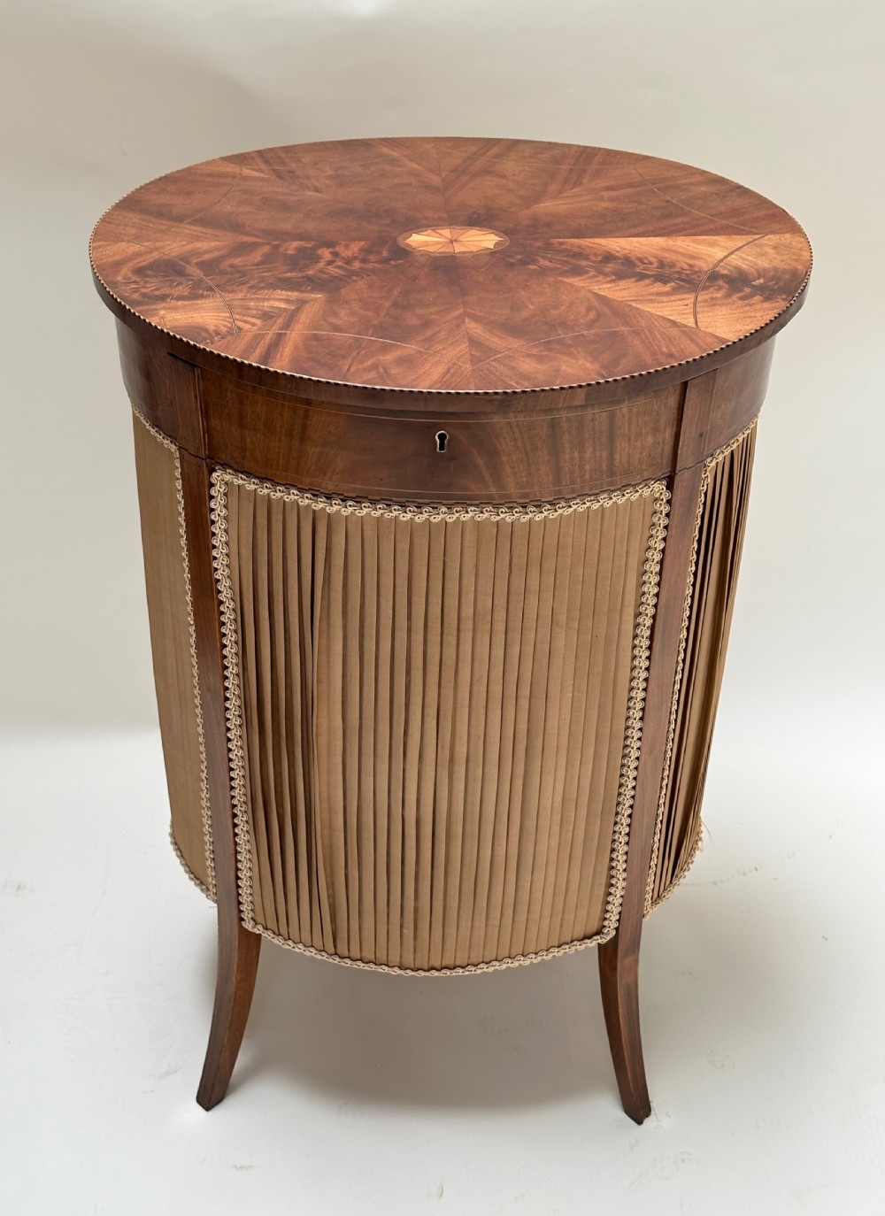 A round antique sewing table featuring a radial walnut veneer top with marquetry inlay, supported by a pleated tan silk basket and slender curved legs. View IV