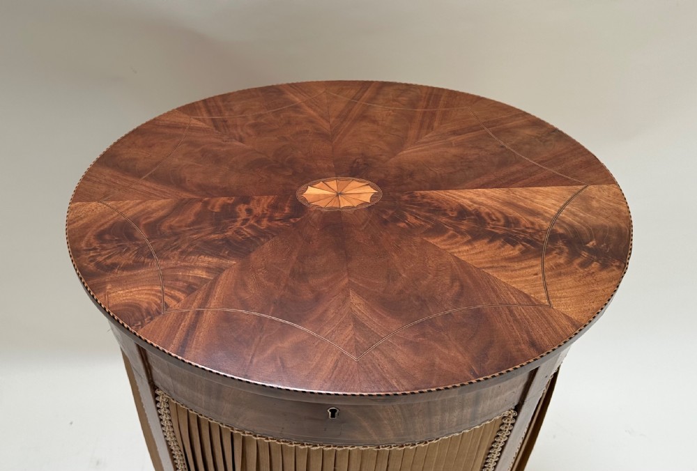 A round antique sewing table featuring a radial walnut veneer top with marquetry inlay, supported by a pleated tan silk basket and slender curved legs. View V