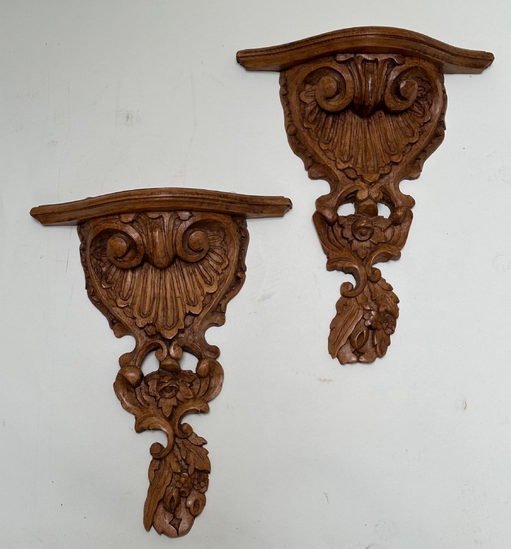 A matching pair of hand-carved warm brown wooden wall brackets featuring ornate scrollwork, shell fluting, and floral motifs.