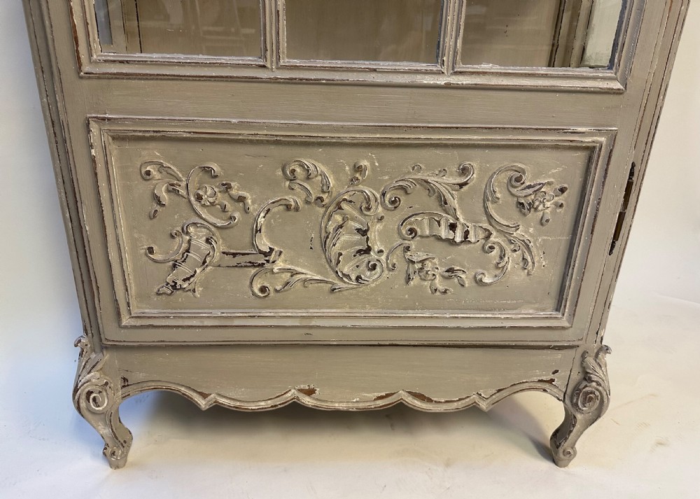 A tall French provincial glazed cabinet featuring a distressed grey painted finish, multi-pane glass doors, intricate scroll carving at the base, and short cabriole legs. View V