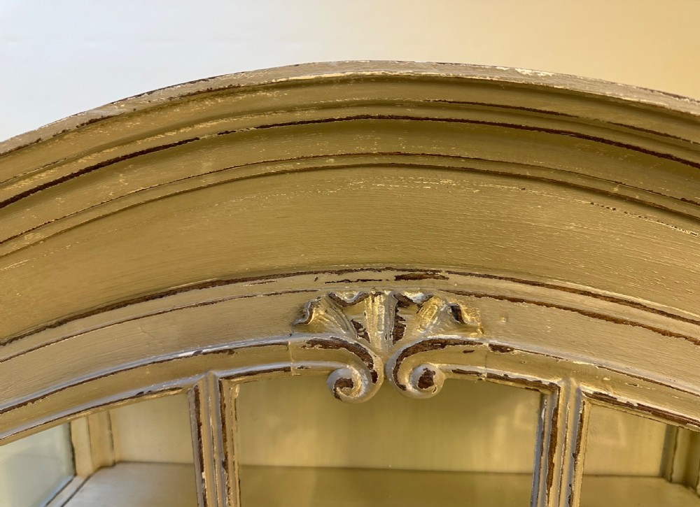 A tall French provincial glazed cabinet featuring a distressed grey painted finish, multi-pane glass doors, intricate scroll carving at the base, and short cabriole legs. View VI