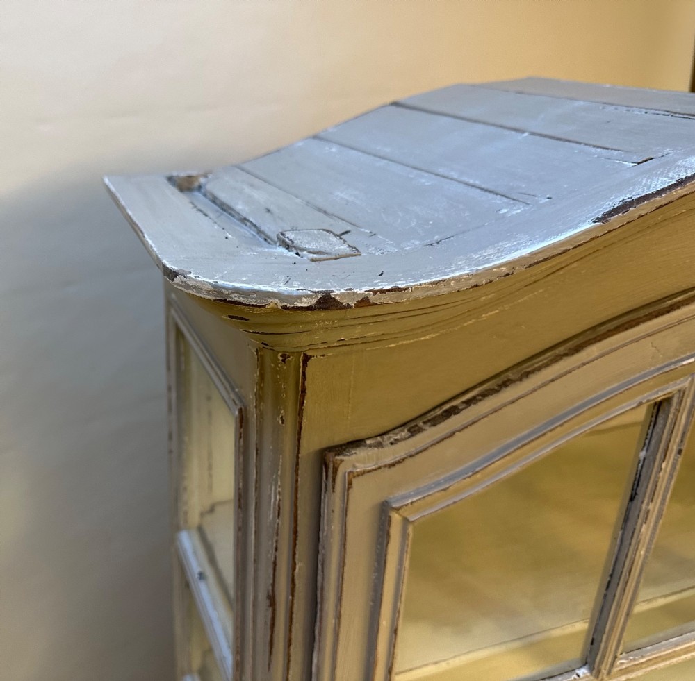 A tall French provincial glazed cabinet featuring a distressed grey painted finish, multi-pane glass doors, intricate scroll carving at the base, and short cabriole legs. View VII