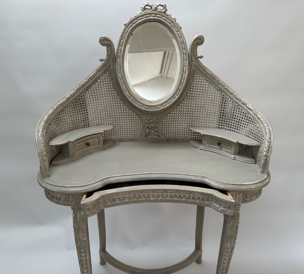 A grey painted French style dressing table with a cane back panel, oval top mirror, small side drawers, and slender carved legs. View V