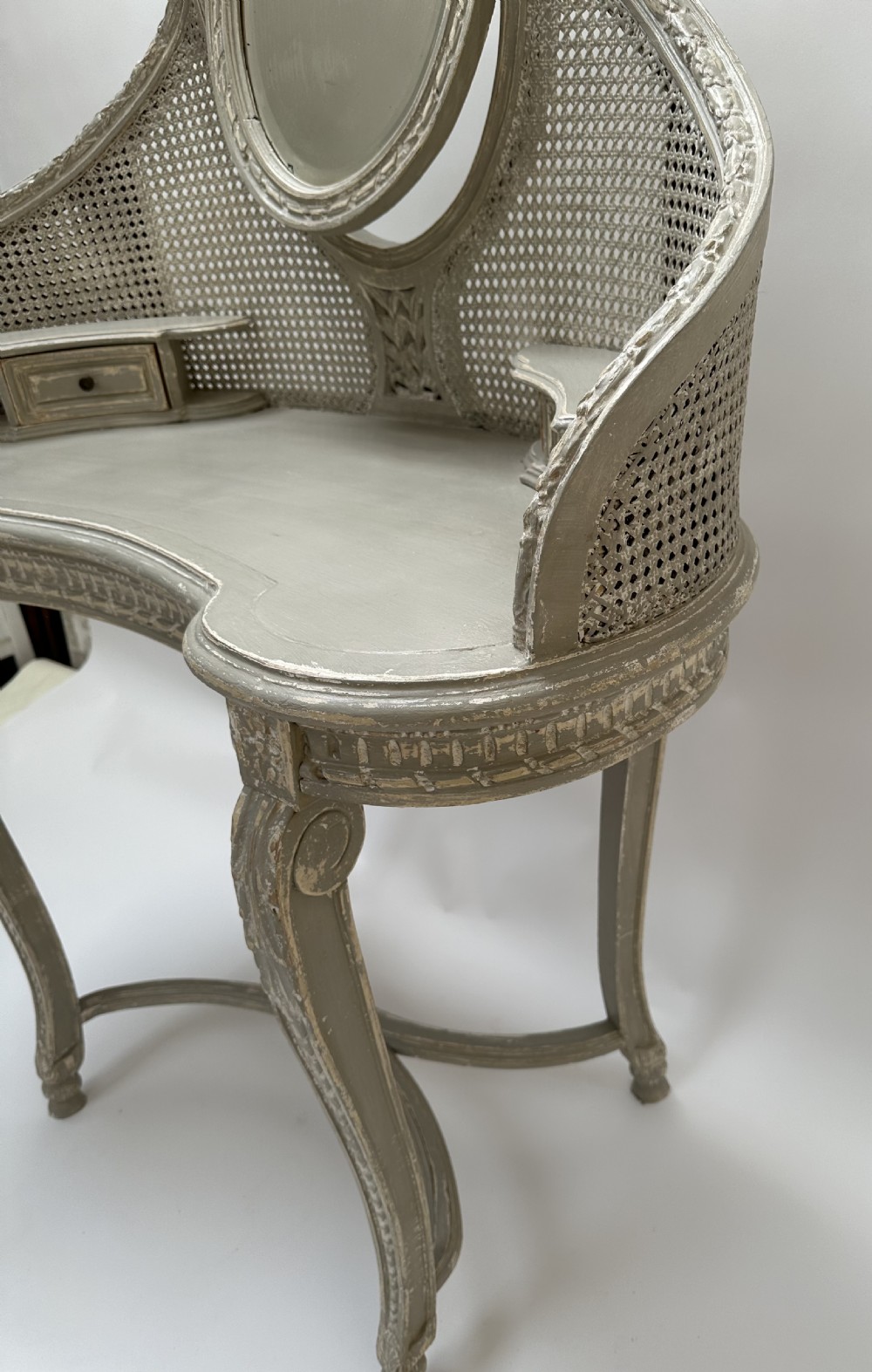 A grey painted French style dressing table with a cane back panel, oval top mirror, small side drawers, and slender carved legs. View VII