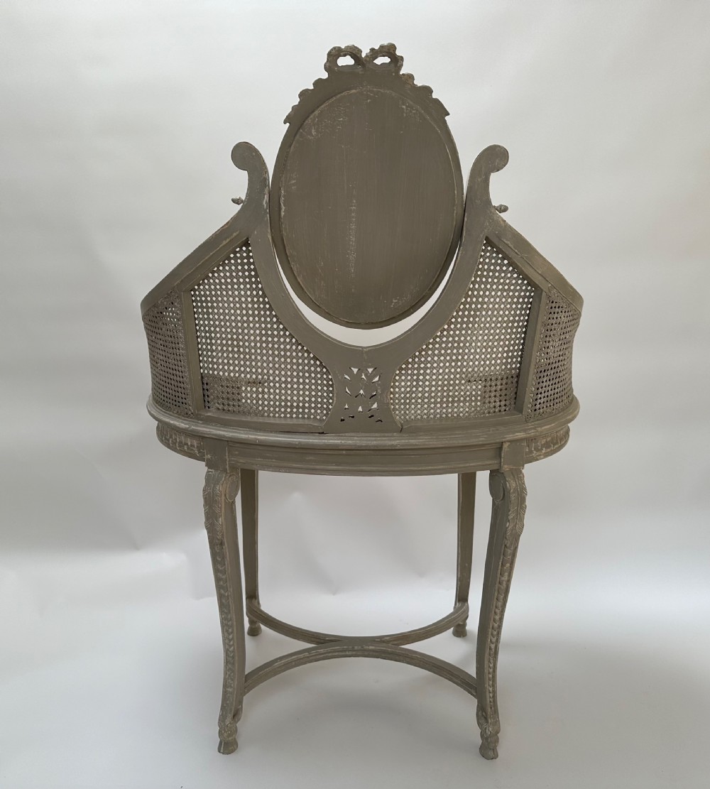 A grey painted French style dressing table with a cane back panel, oval top mirror, small side drawers, and slender carved legs. View VIII