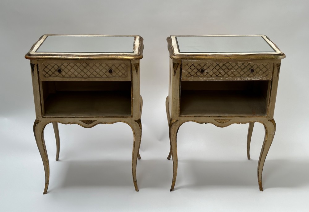 A pair of cream and gold Florentine bedside tables featuring mirrored tops, open lower compartments, carved lattice drawers, and slender cabriole legs.