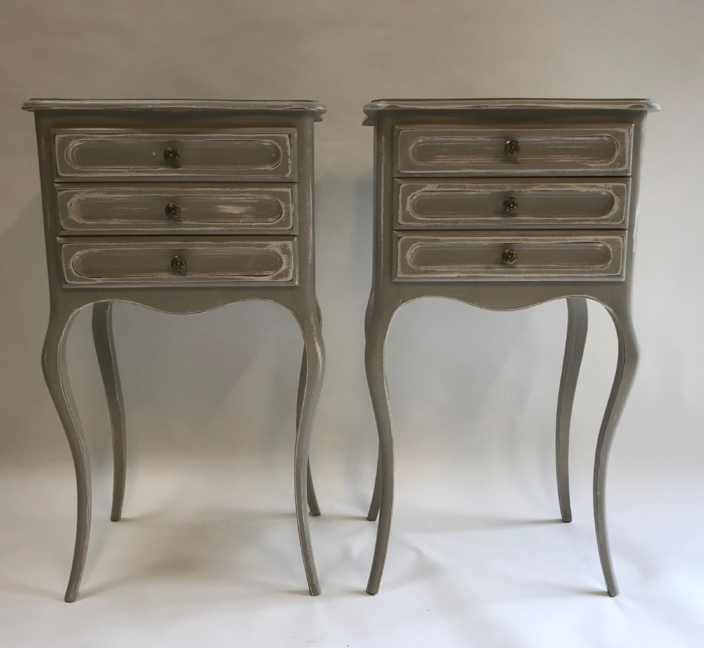 A pair of slender grey painted French provincial bedside tables featuring three drawers and tall cabriole legs.