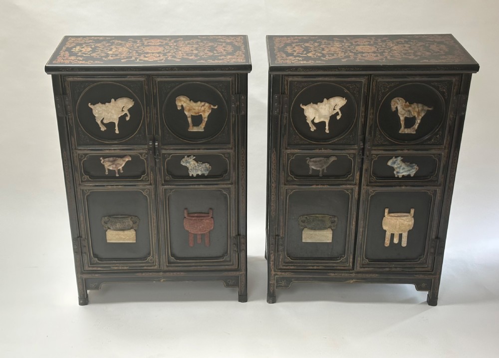 A pair of black lacquered two-door Chinoiserie cabinets featuring carved hardstone inlays of horses, animals, and vessels, topped with gilt scrollwork motifs.